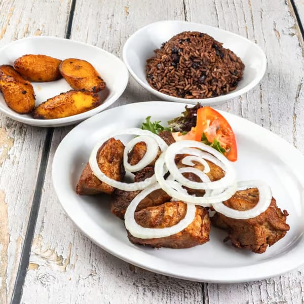 Cuban fried pork chunks with onions, rice and beans, and fried plantains on a white wooden table.