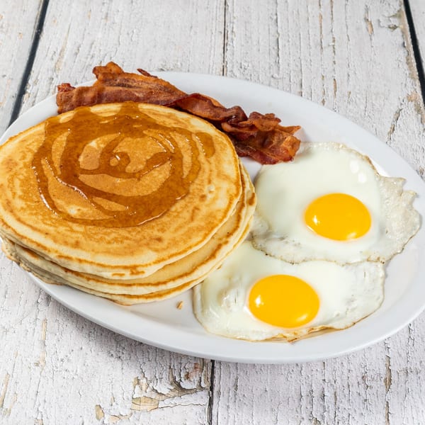 A breakfast plate with a stack of pancakes drizzled with syrup, crispy bacon, and two fried eggs.