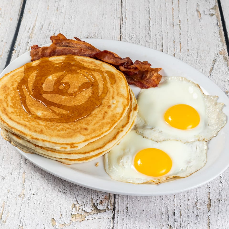 A breakfast plate with a stack of pancakes drizzled with syrup, crispy bacon, and two fried eggs.