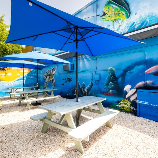 Outdoor seating area with blue umbrellas, picnic tables on gravel, and a vibrant ocean-themed mural.