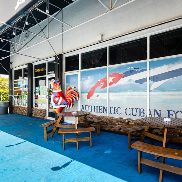Dennys Latin Cafe exterior with blue patio, picnic tables, and a large Cuban flag window decal.